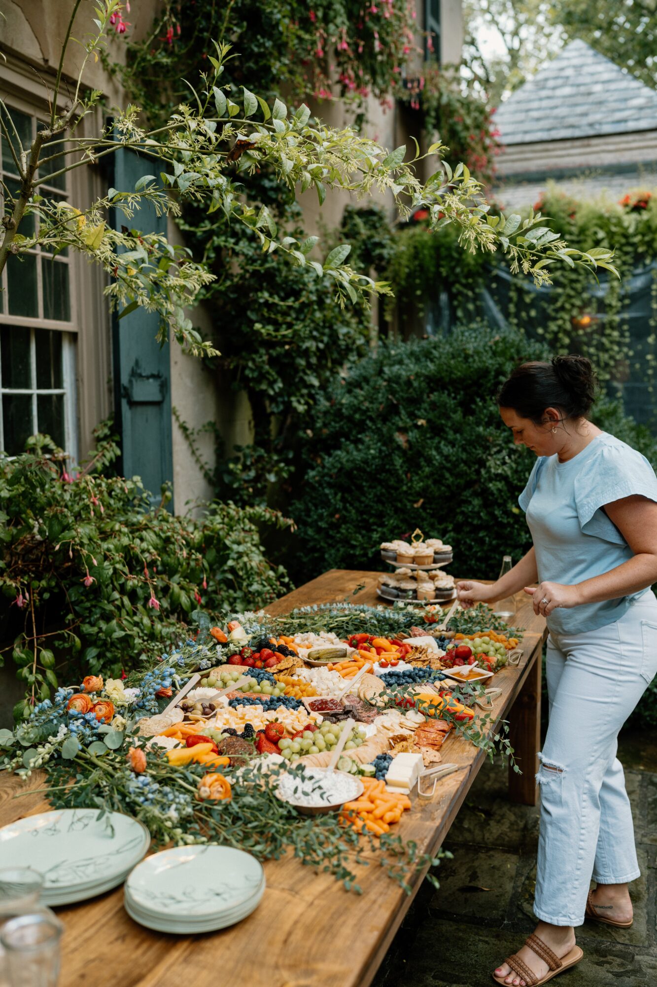 Woman in light blue shirt and jeans selecting food from a table outdoors, surrounded by greenery and flowers.