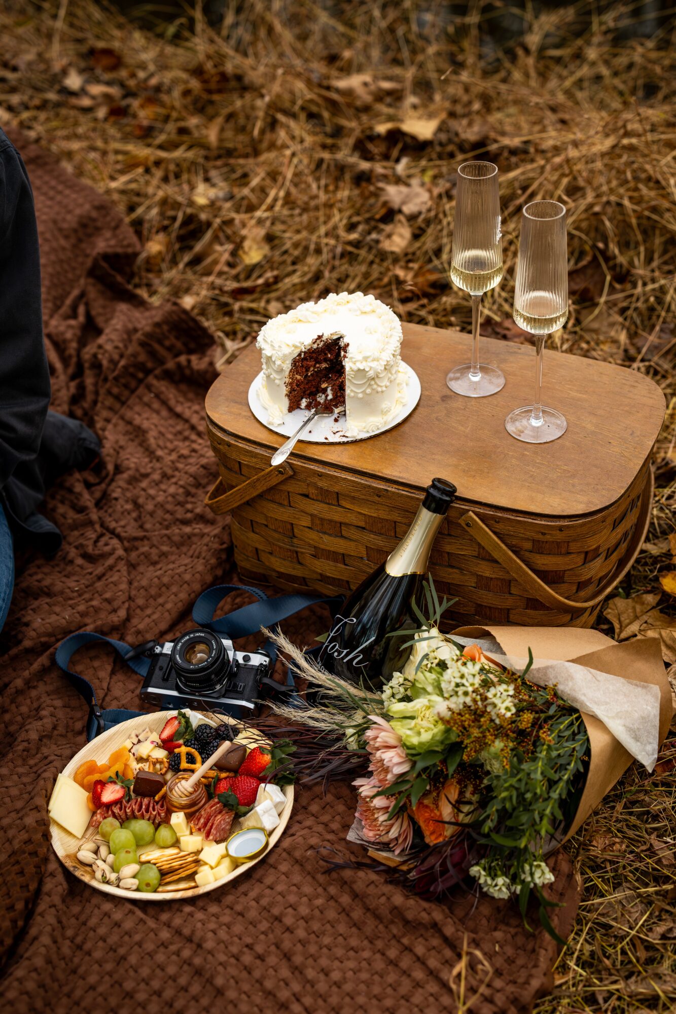 A cake with white frosting and a slice removed, two champagne glasses, a bottle of champagne, a plate of fruit, and a bouquet on a picnic blanket outdoors.