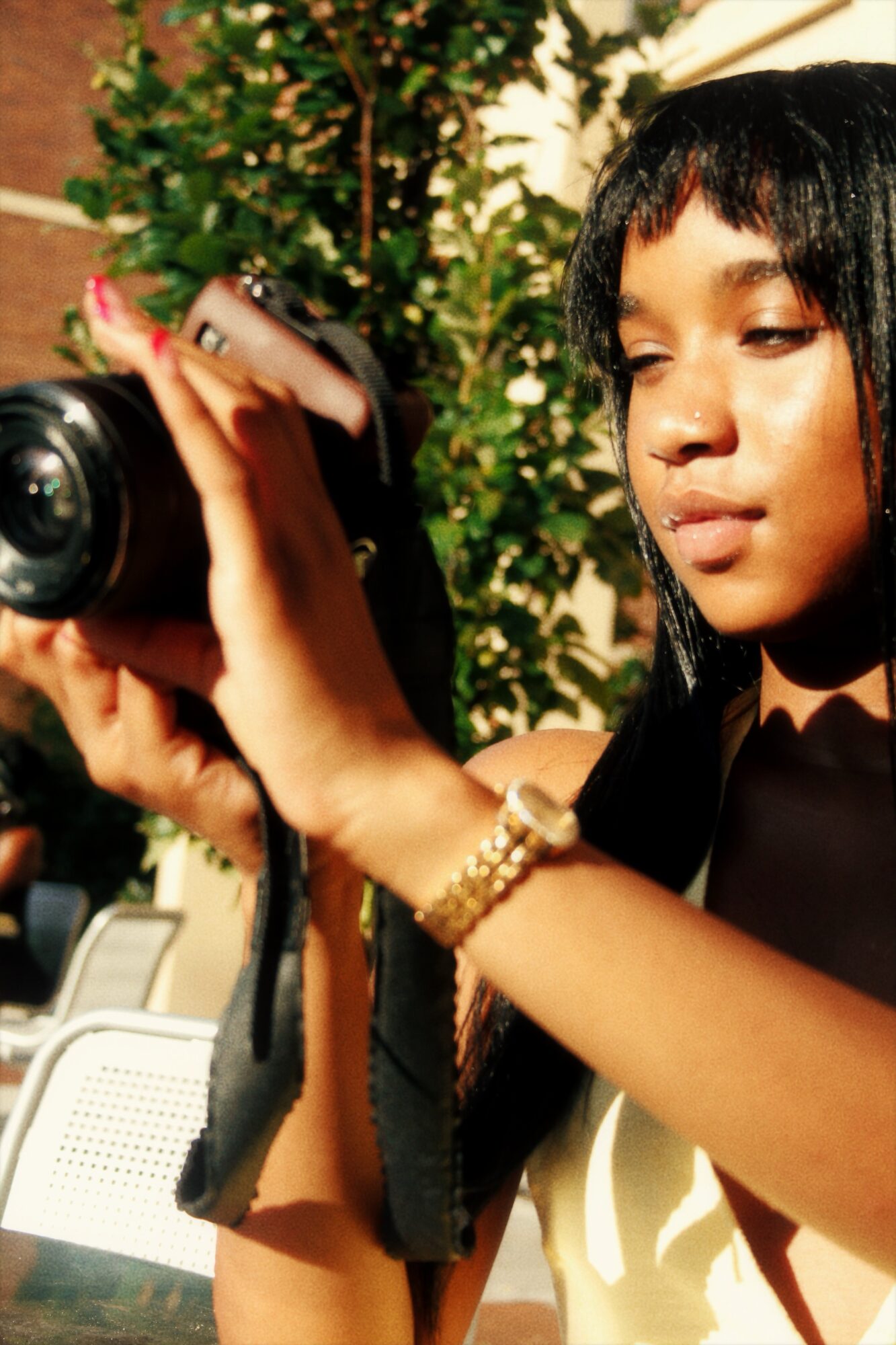 Young woman with dark hair and a gold watch holds a camera outdoors, with greenery in the background.