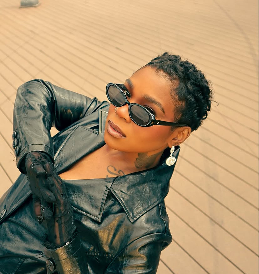 Woman with short curly hair wearing sunglasses, leather jacket, and gloves, standing on wooden deck.