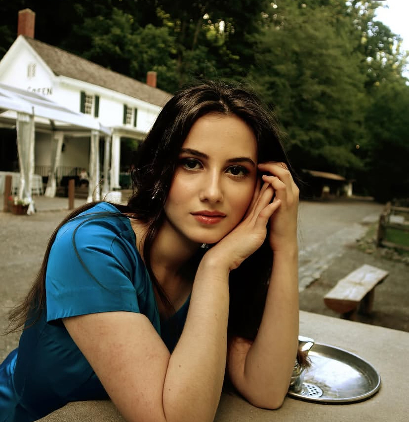 Young woman with long dark hair resting her face on her hand outdoors, wearing a blue shirt, with a building and trees in background.