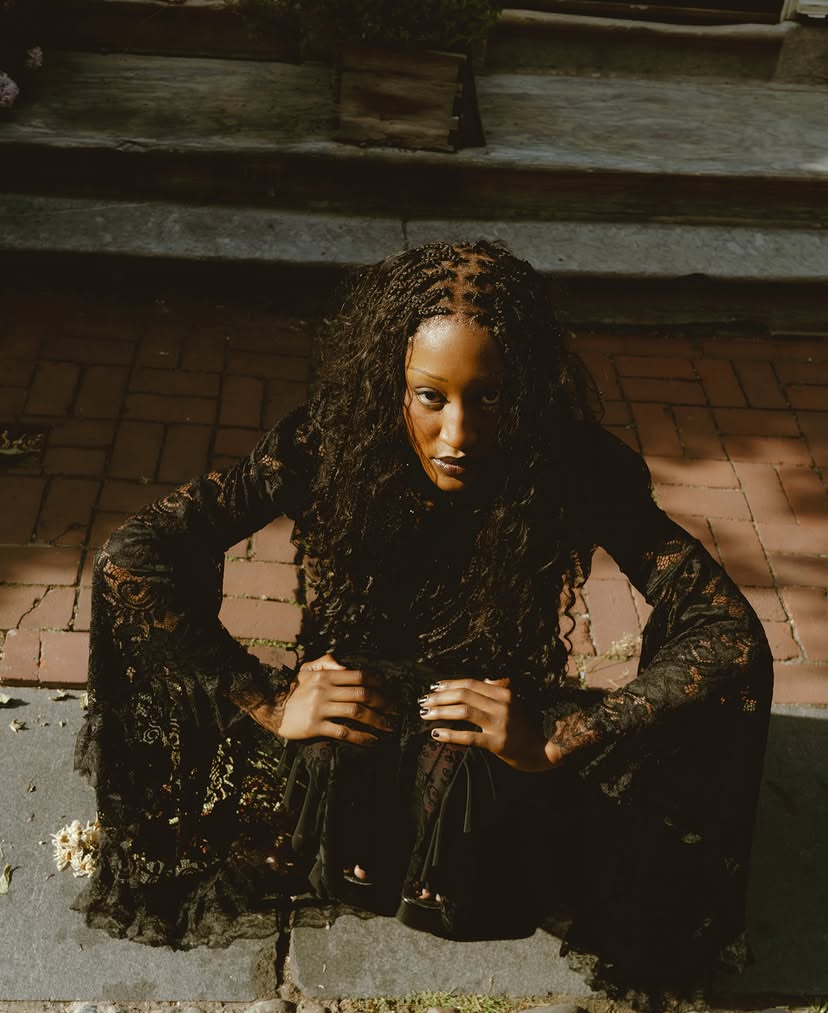 Woman with curly hair crouching outdoors at night, wearing a black lace dress, hands on hips, looking at camera.