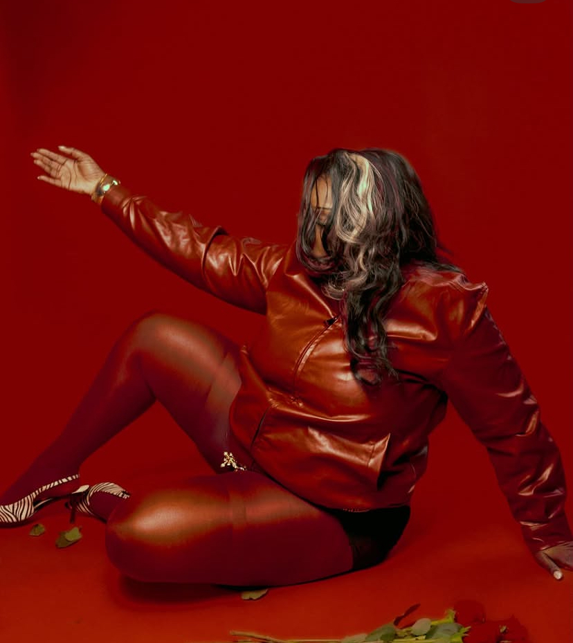 Person sitting on the floor with one arm extended, wearing a leather jacket and tights, against a red background.