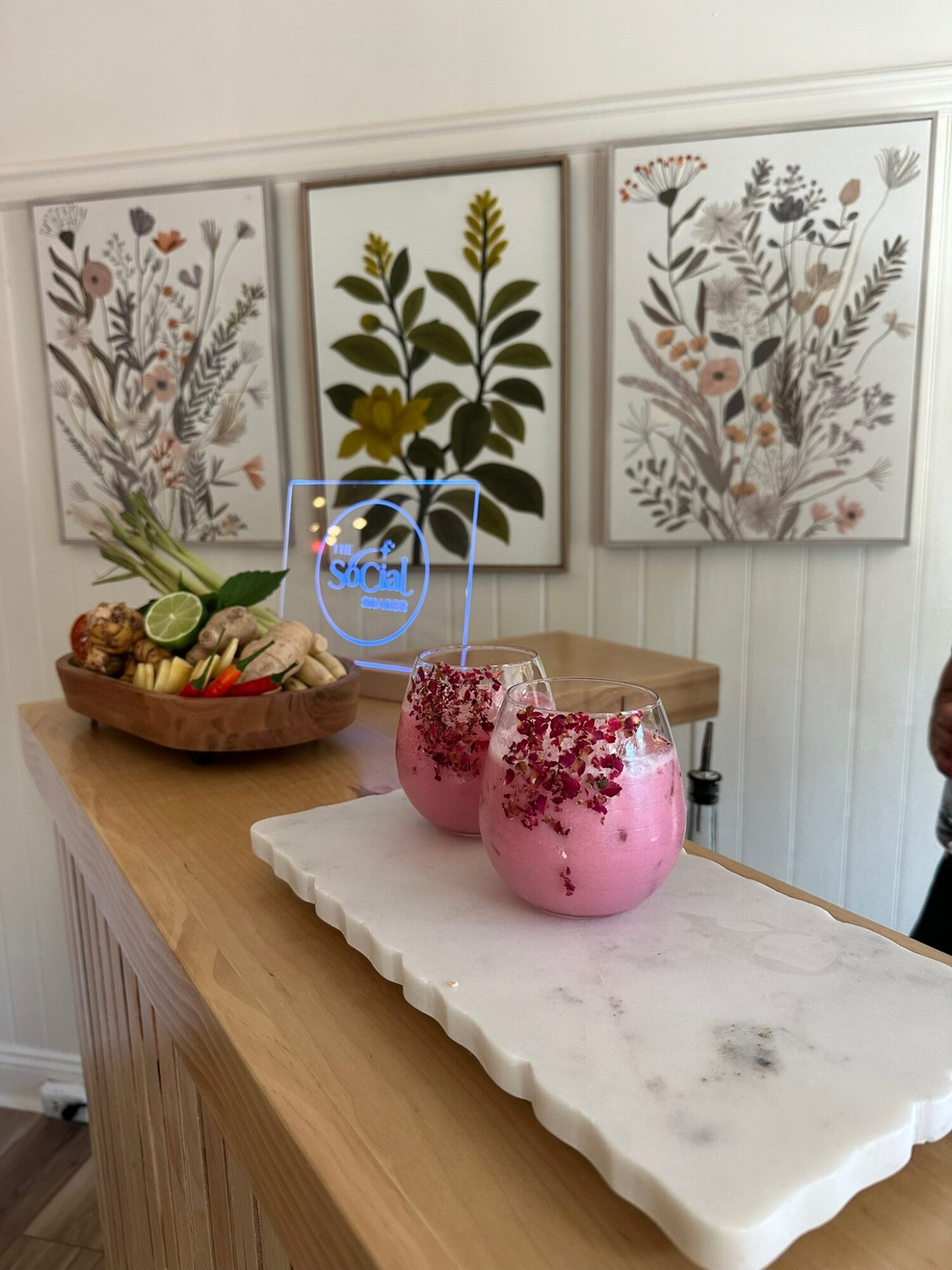 Three pink candles with floral designs on a white tray, a wooden bowl with vegetables and a plant, and three botanical prints on the wall.