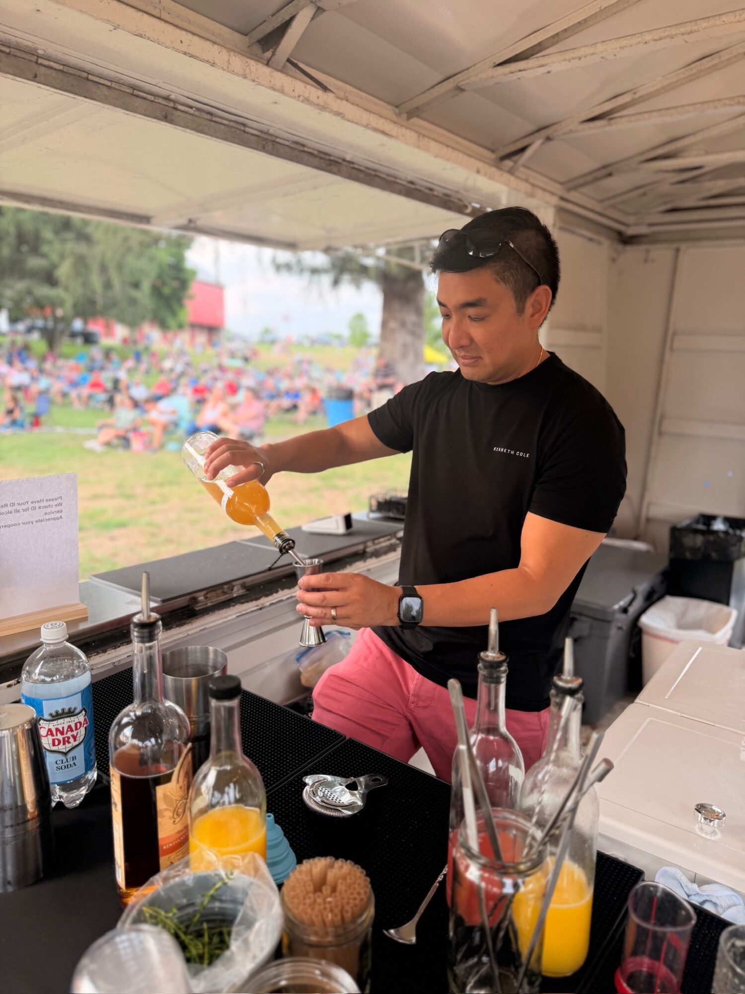 Man pouring drink in a bar with bottles and glasses, window showing outdoor scene with people and trees