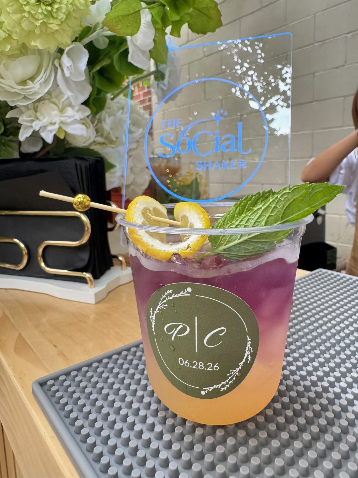Colorful layered drink with lemon slices, mint, and a leaf, on a gray mat with a floral arrangement in background.
