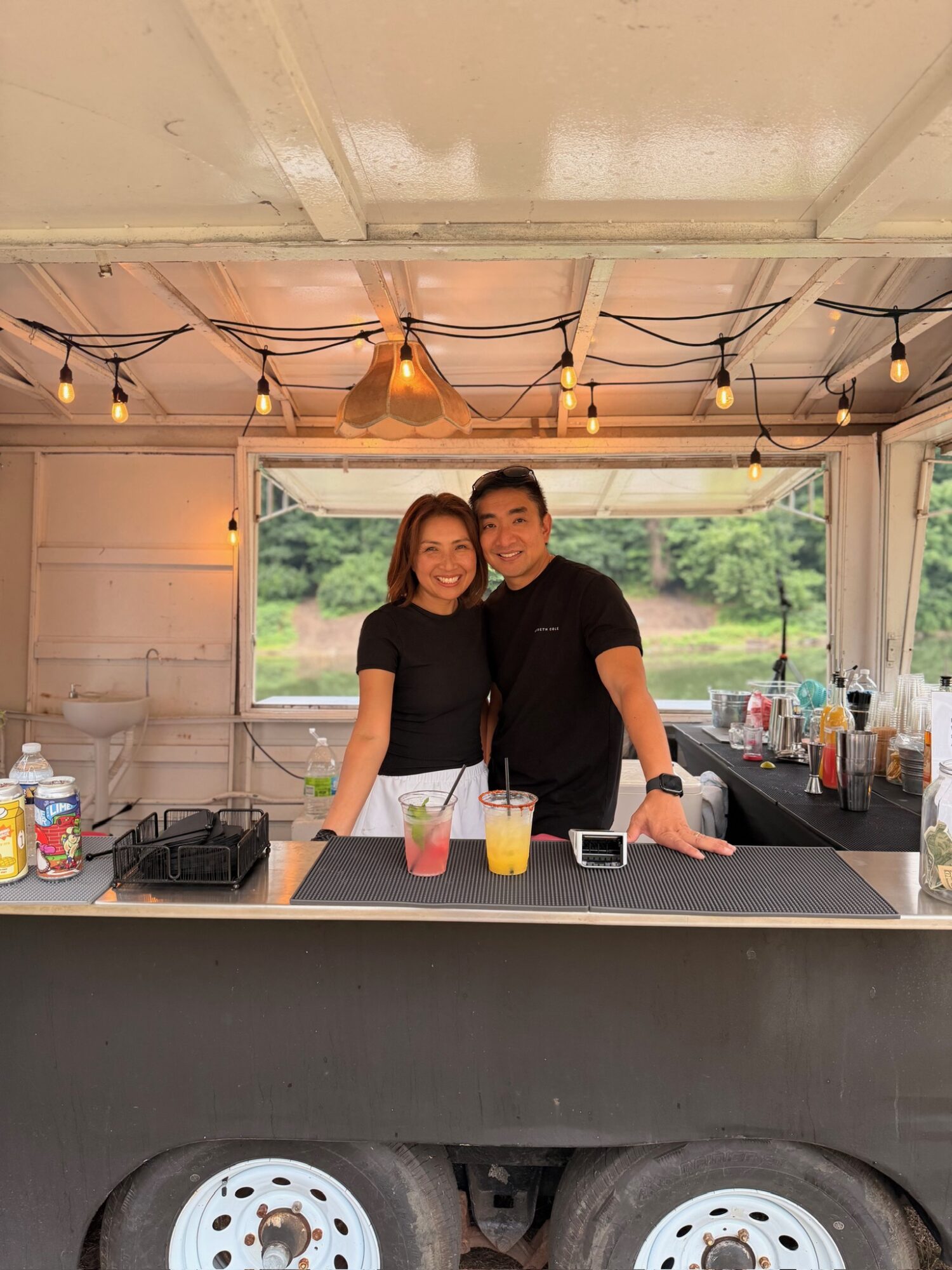 Two people stand behind a bar counter inside a tent, smiling, with drinks and bar supplies visible, outdoors in the background.