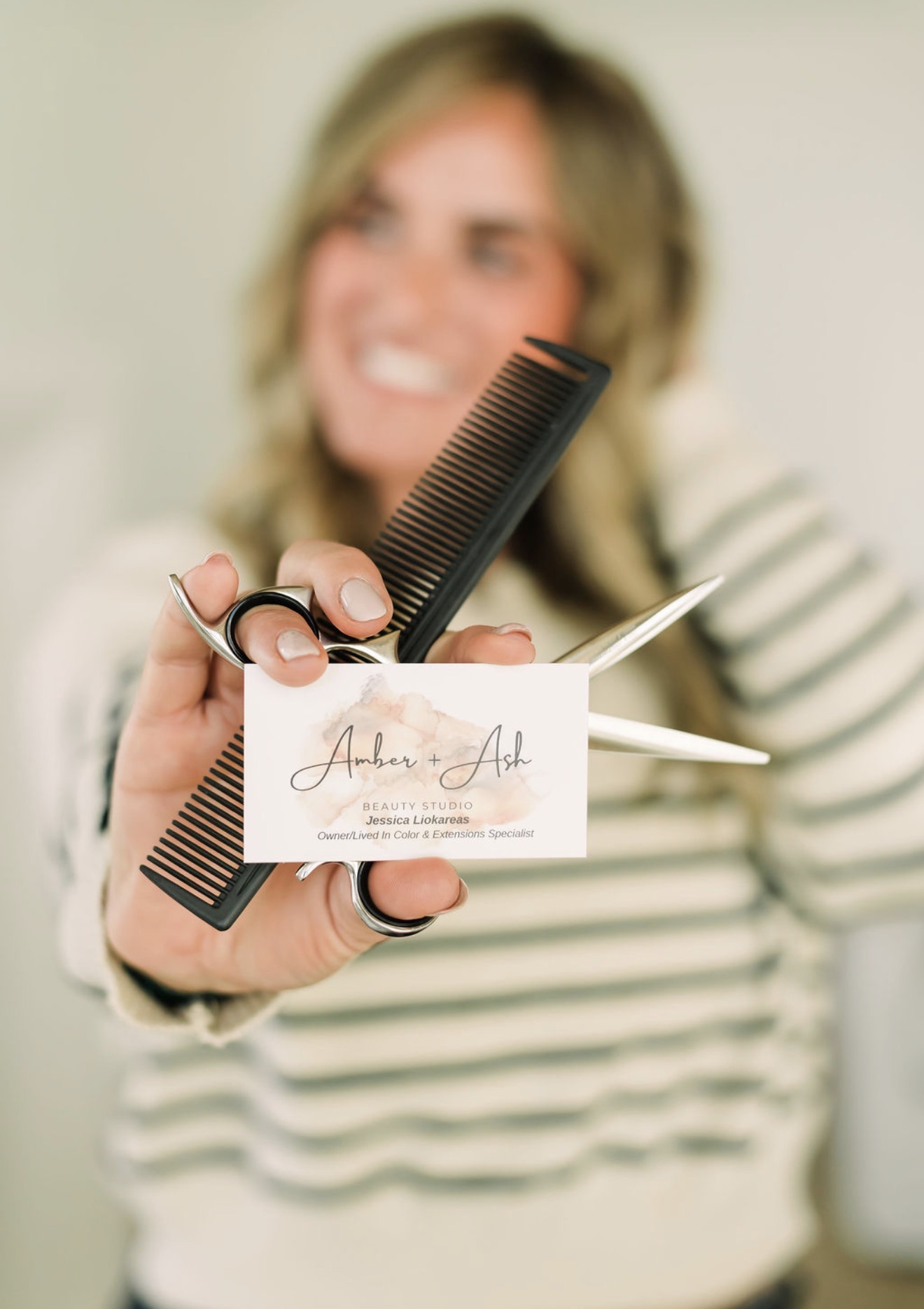 Woman with blonde hair holding a black comb and business card, smiling, wearing a striped shirt.