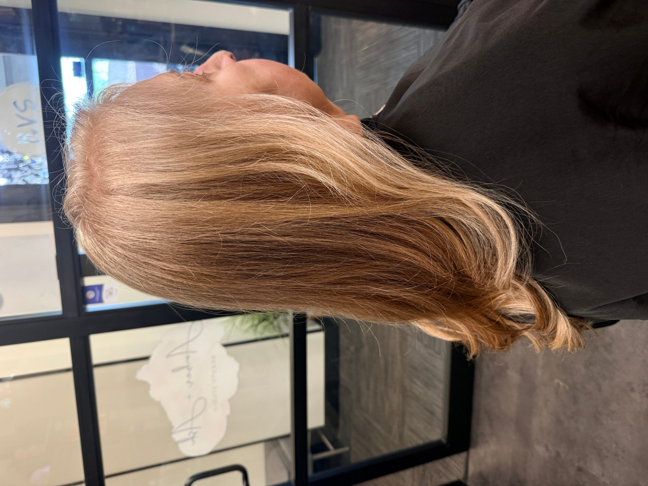 Person with long, blonde hair, viewed from the side, sitting indoors near a window.