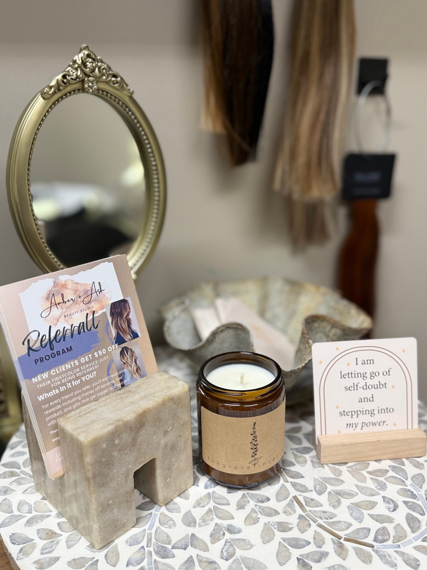 Decorative mirror, candle, and motivational card on a patterned surface, with hair samples hanging in the background.