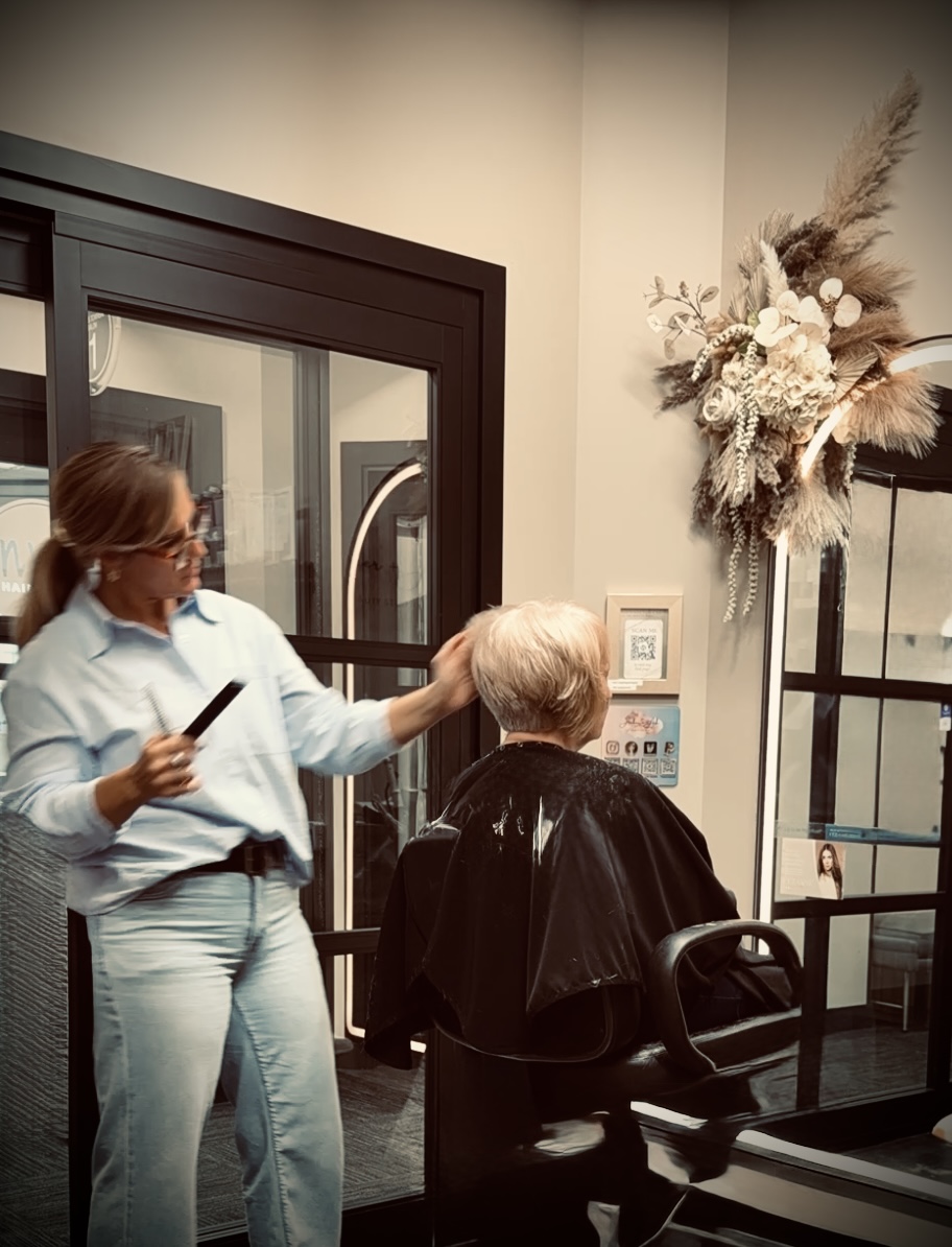 Person with short hair getting a haircut in a salon, with a stylist holding scissors and a mirror, and a decorative floral arrangement on the wall.
