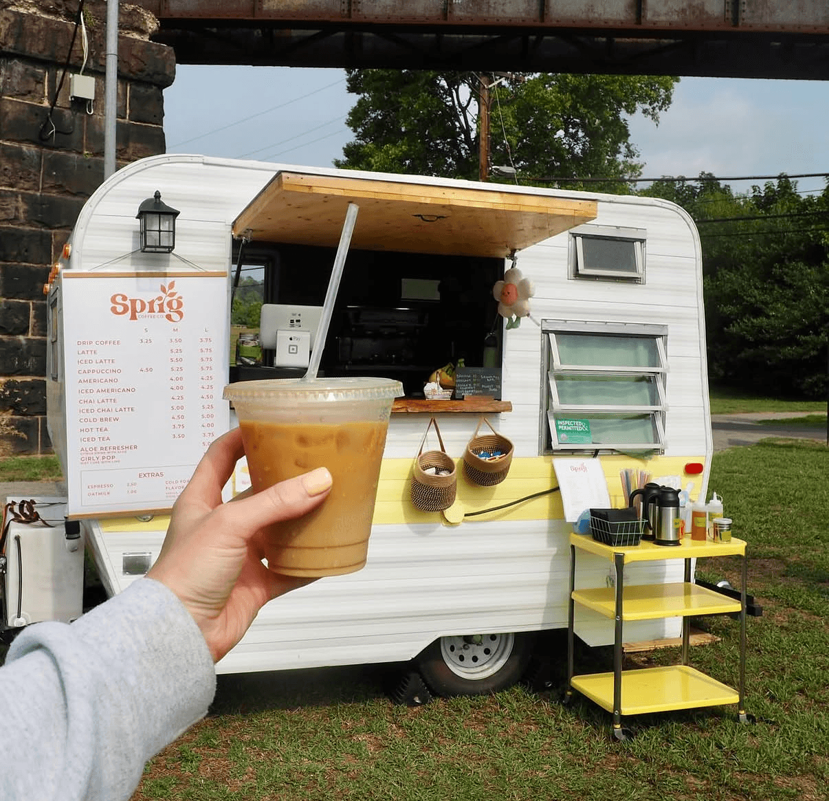Person holding iced coffee in front of a food truck with a menu and yellow cart outside.