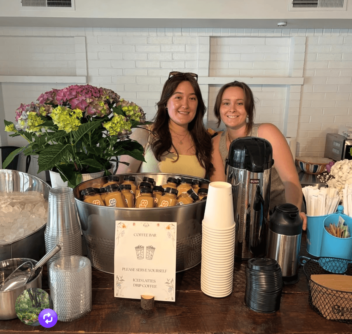 Two women smiling behind a coffee station with flowers, cups, and coffee equipment in a cafe setting.