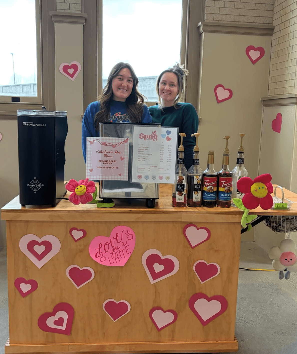 Two women stand behind a decorated table with pink hearts, drinks, and a menu, in a room with windows and brick walls.
