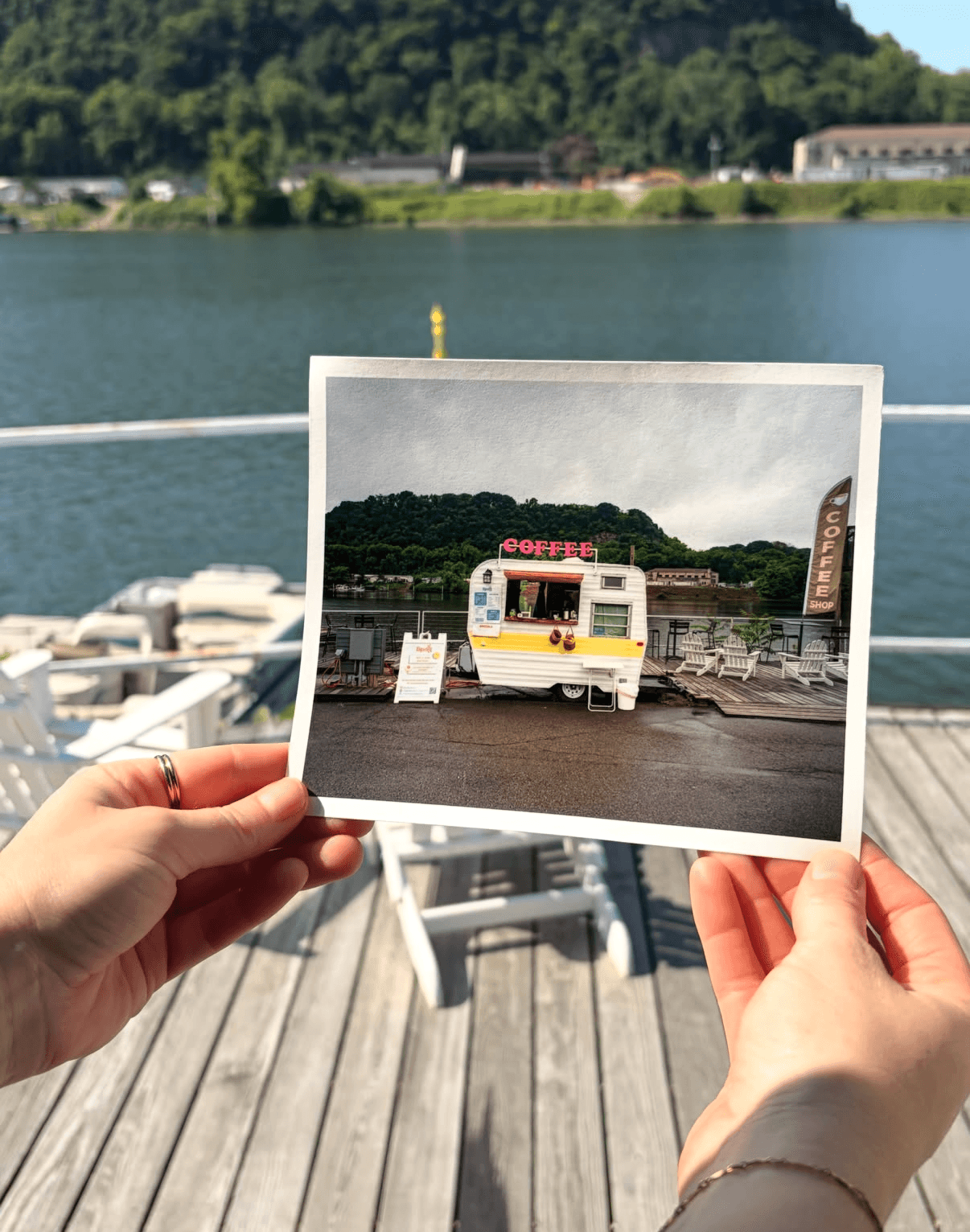 Person holding a photo of a yellow and white bus in front of a body of water and green hills.