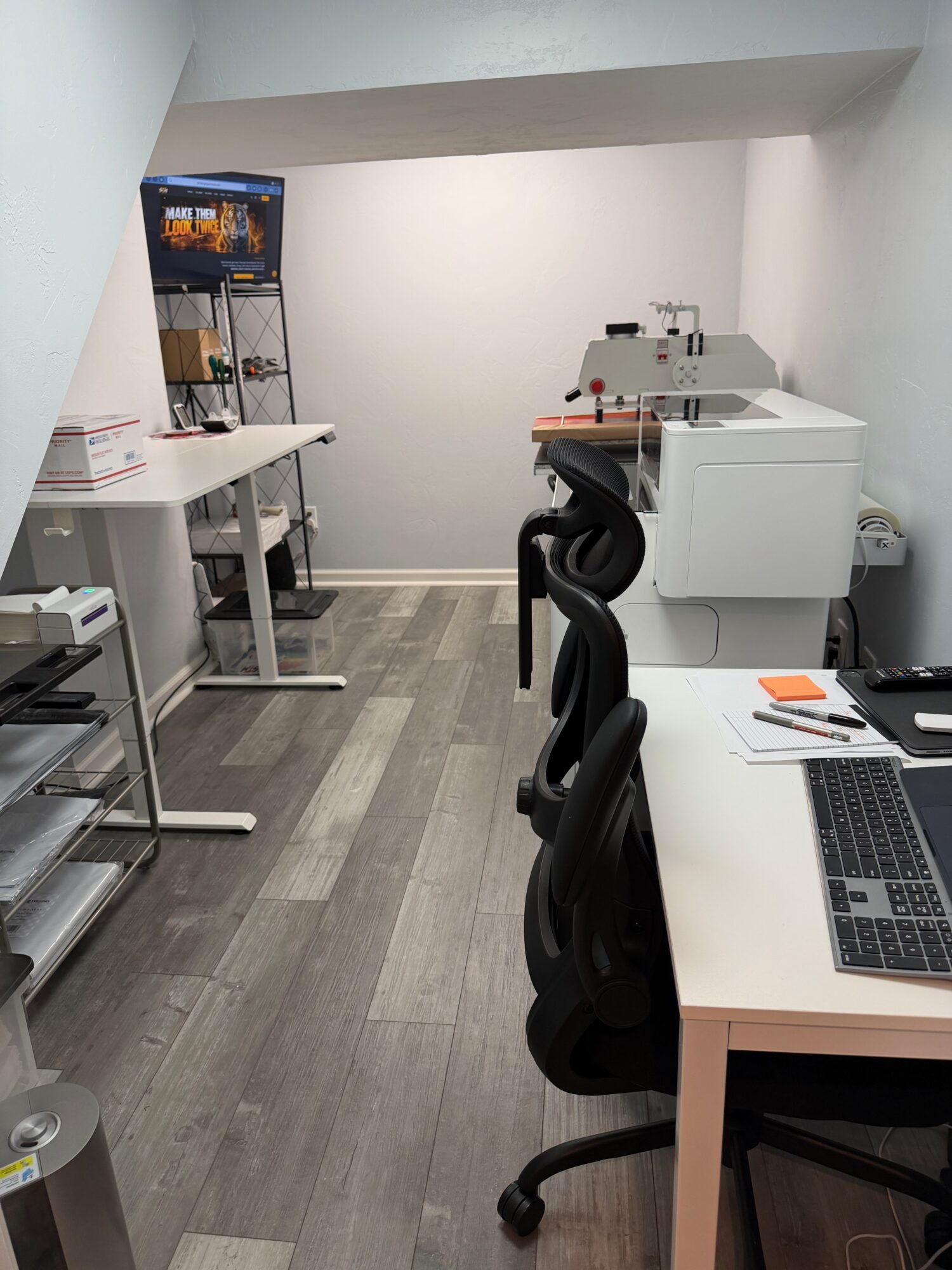 Office space with desks, chairs, a laptop, and a machine on a white table, with a white wall and gray flooring.