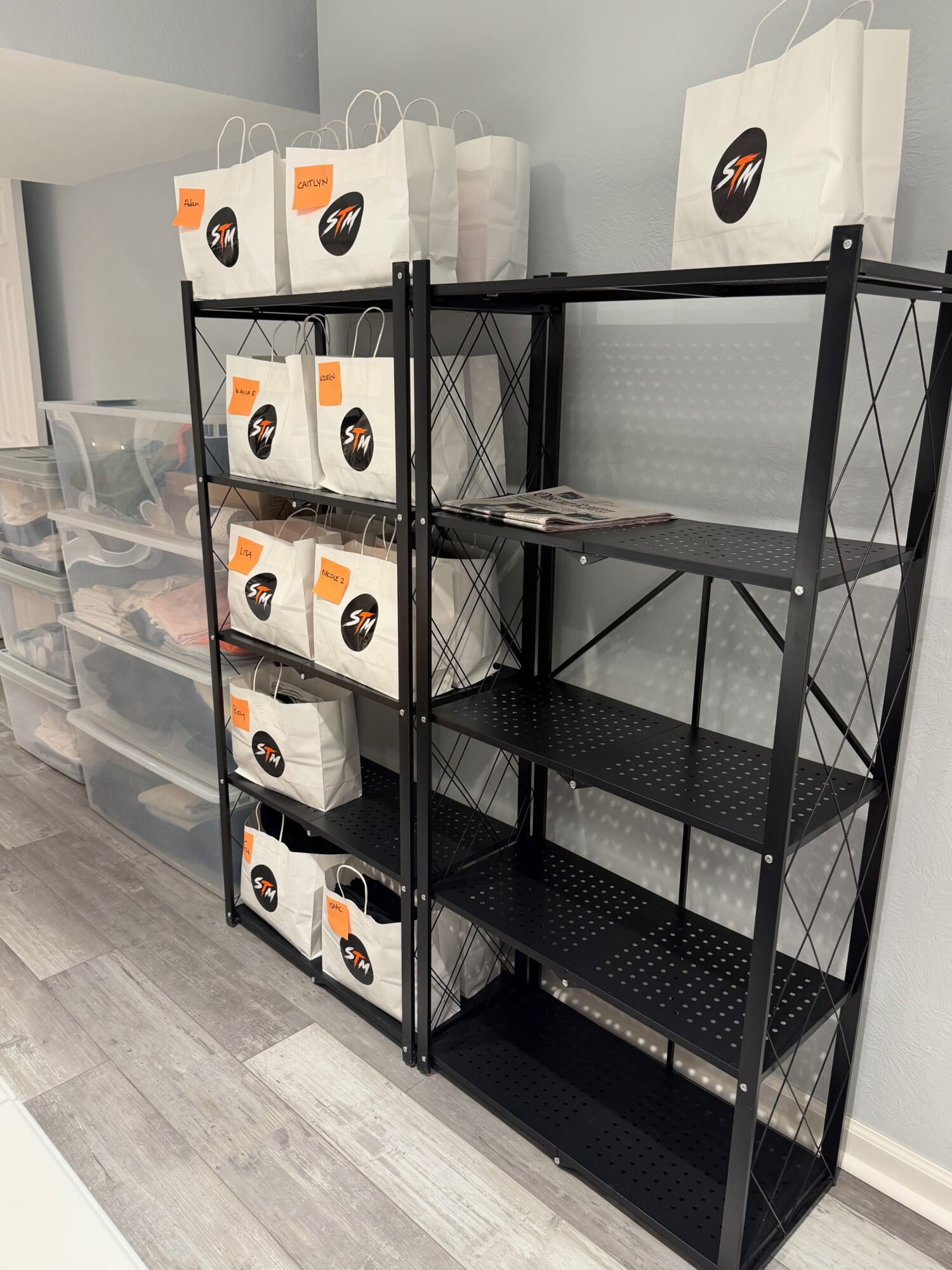 Two black metal shelving units against a wall, with white shopping bags on top and plastic storage drawers nearby.