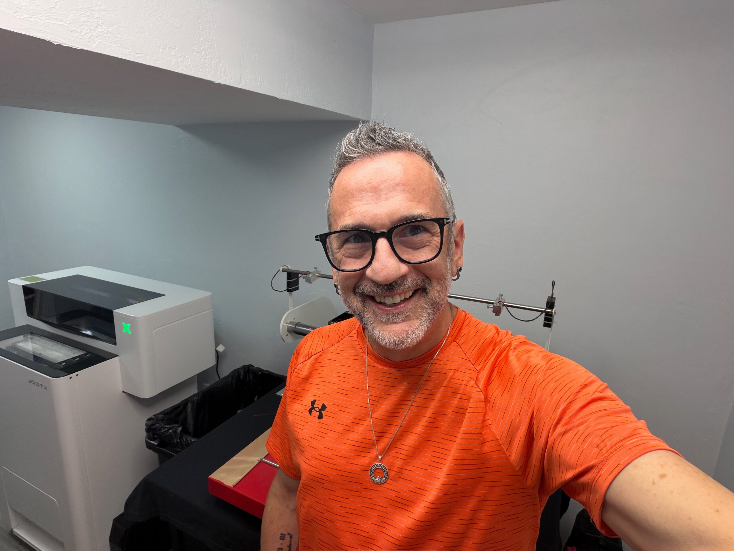 Smiling man with glasses and gray hair wearing an orange shirt, taking a selfie indoors with a printer and equipment in the background.