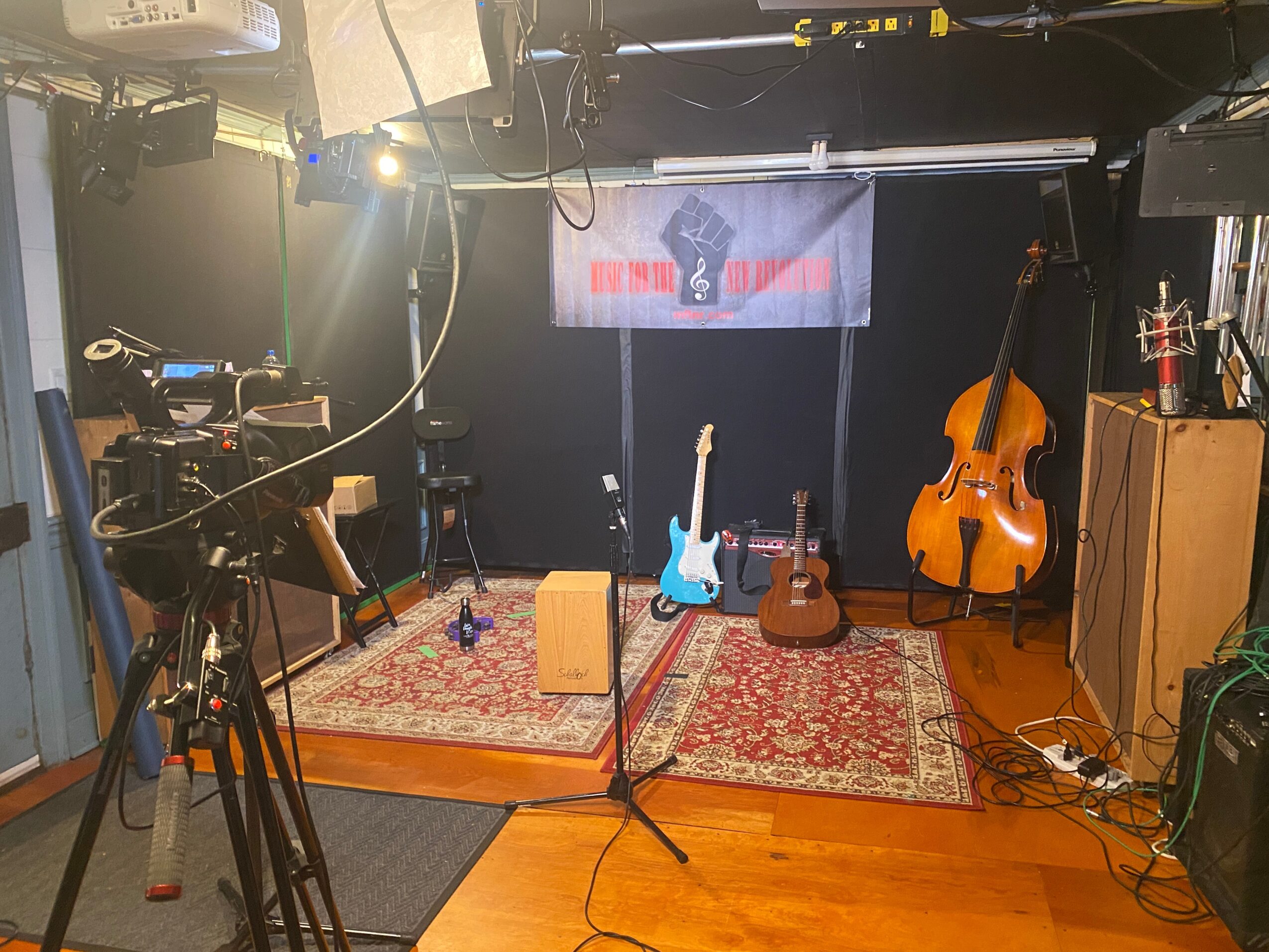 Stage with musical instruments including guitars, a double bass, and a cajón, with a banner and microphones, in a recording studio.