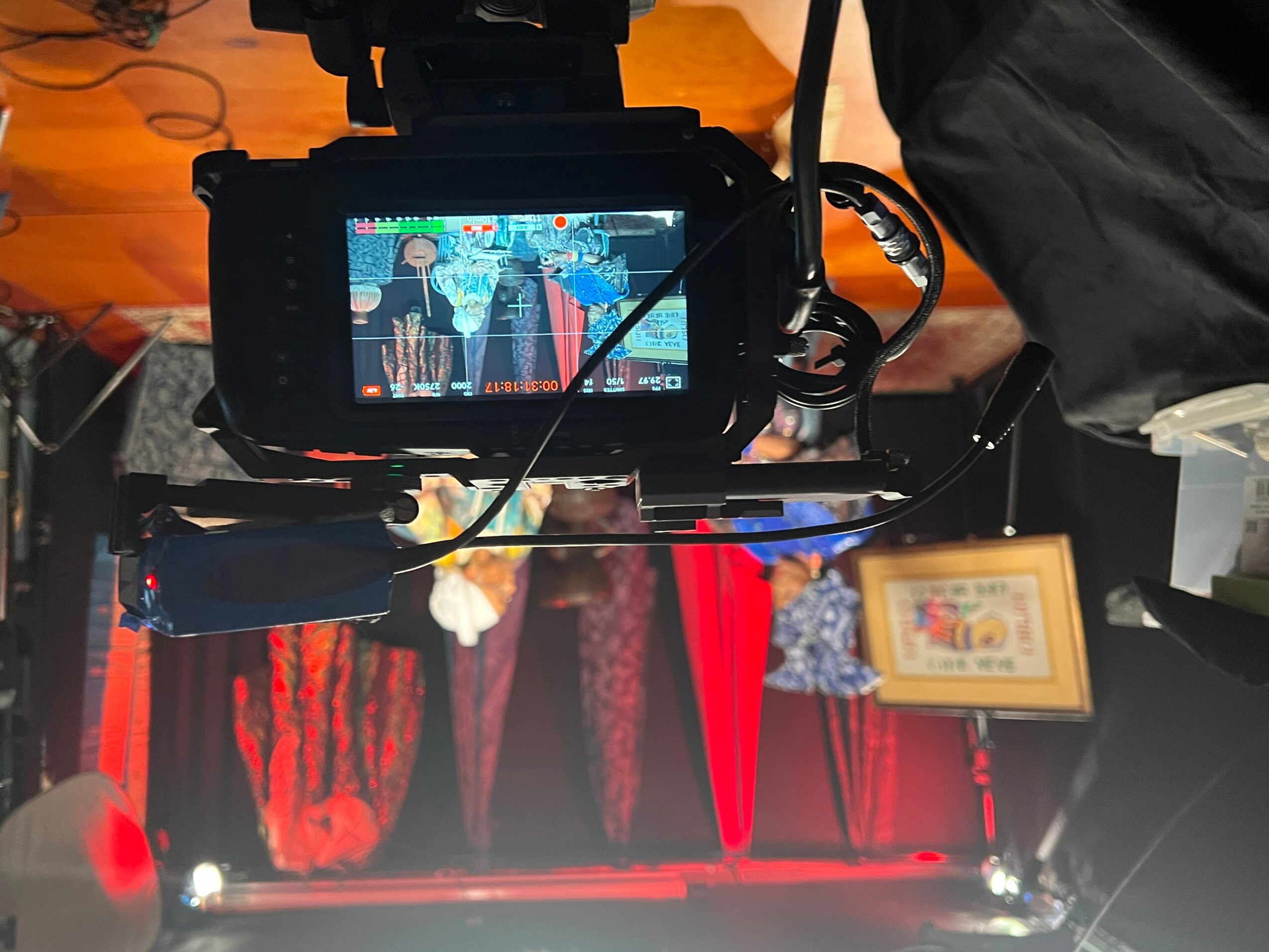 Camera view of a stage with performers, red curtains, and a framed picture, seen from behind the camera.