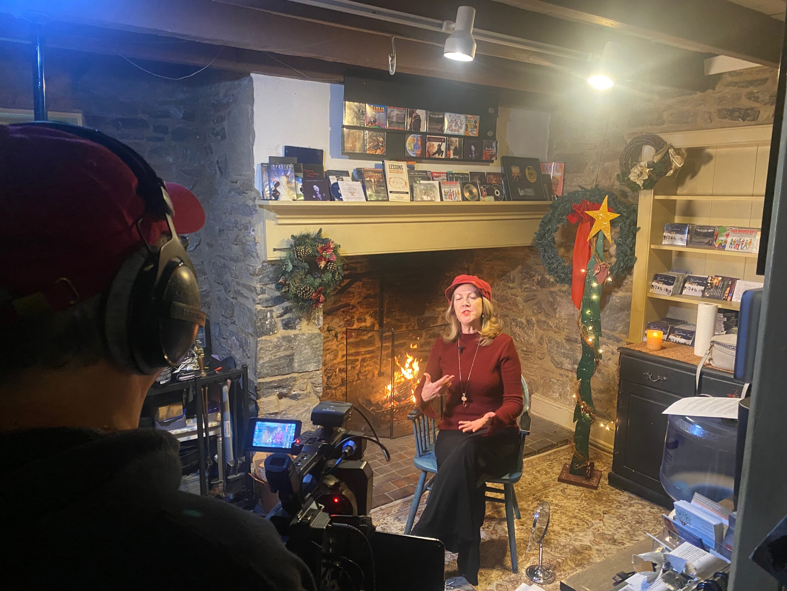 Person with headphones and red cap filming a woman sitting by a fireplace decorated for Christmas, holding a sparkler.