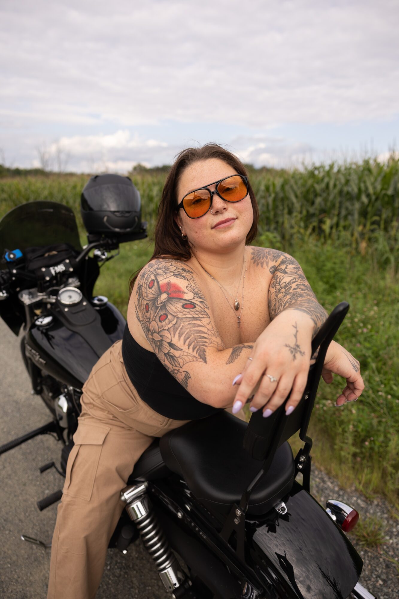 Woman with tattoos and sunglasses sitting on a black motorcycle outdoors, green field and cloudy sky in background.