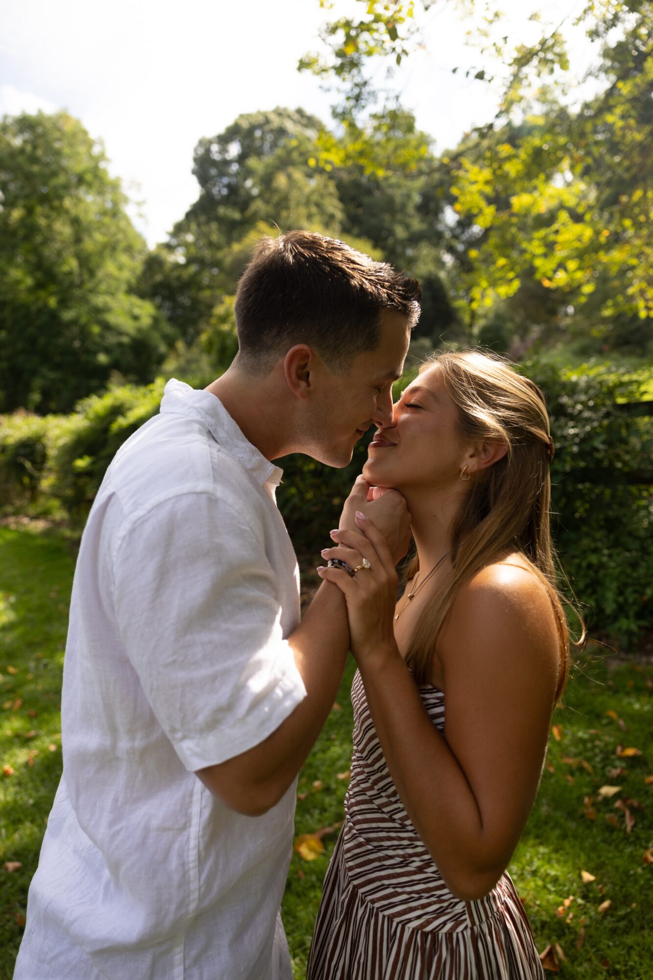 Young couple standing outdoors, about to kiss, surrounded by trees and greenery, sunlight filtering through leaves.