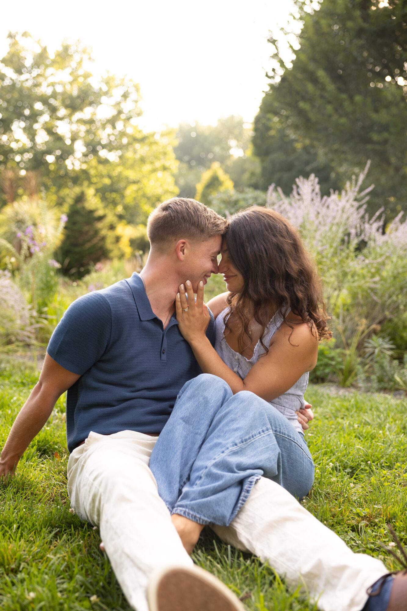 Couple sitting on grass in a garden, touching foreheads, surrounded by greenery and flowers, sunlight filtering through trees.