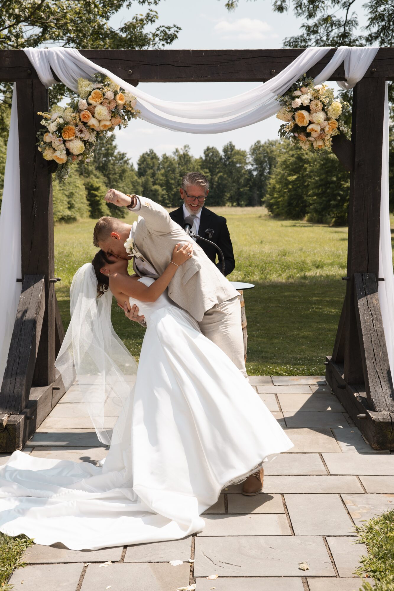 Bride and groom kiss under an arch decorated with flowers outdoors, with trees and grass in the background.