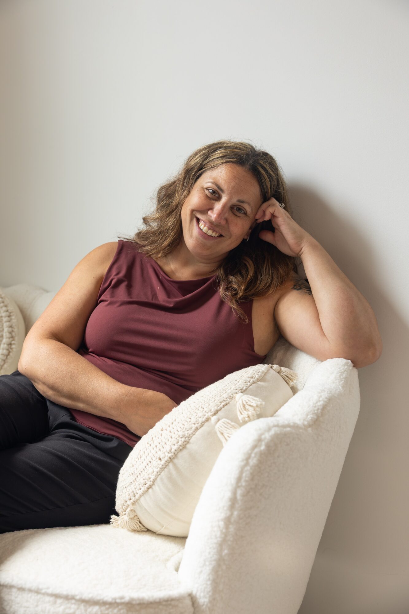Woman with wavy hair smiling, sitting on a white chair, resting her head on her hand, wearing a sleeveless maroon top.