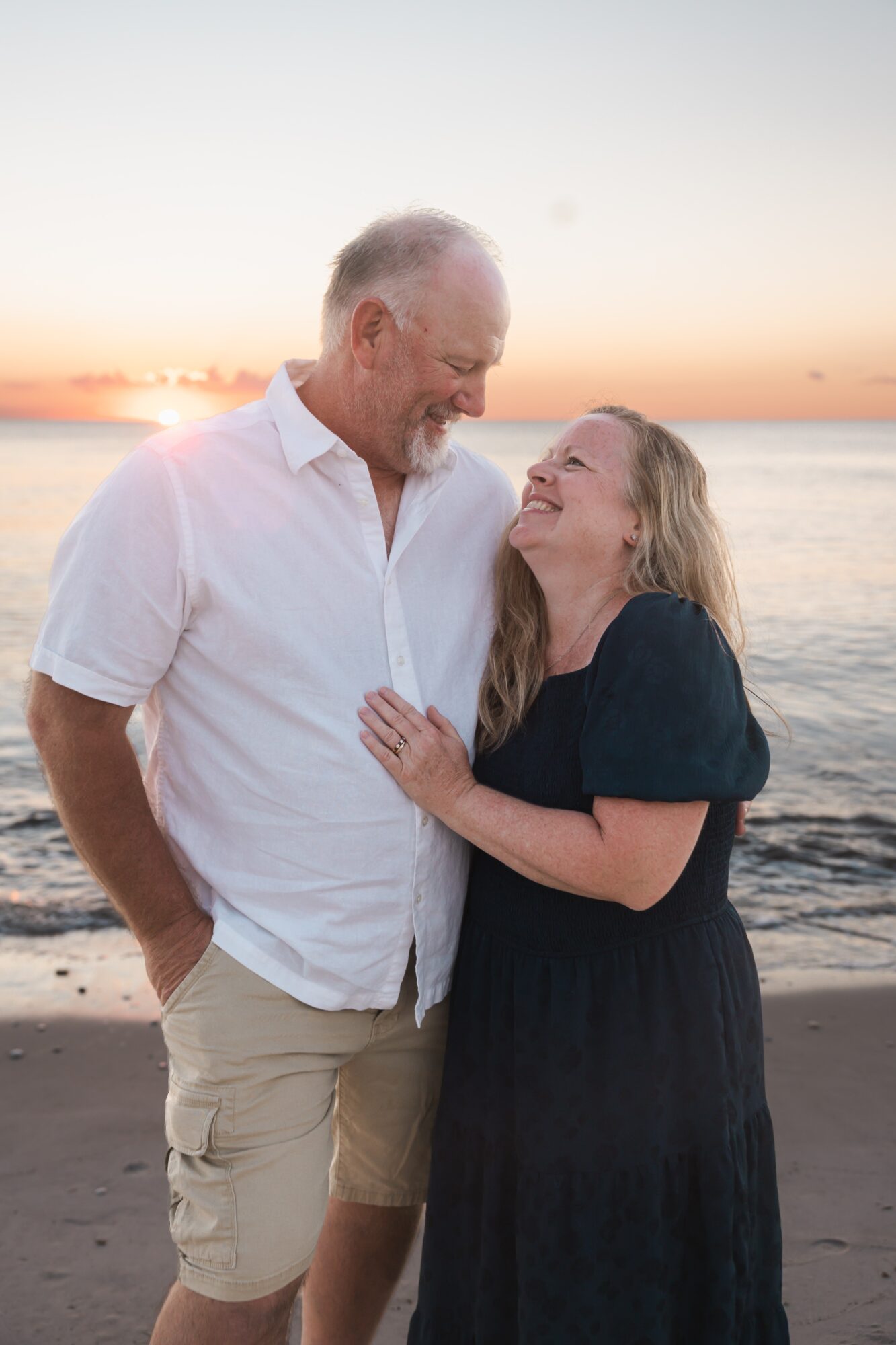 A man and woman smiling and looking at each other on a beach at sunset, with the ocean in the background.