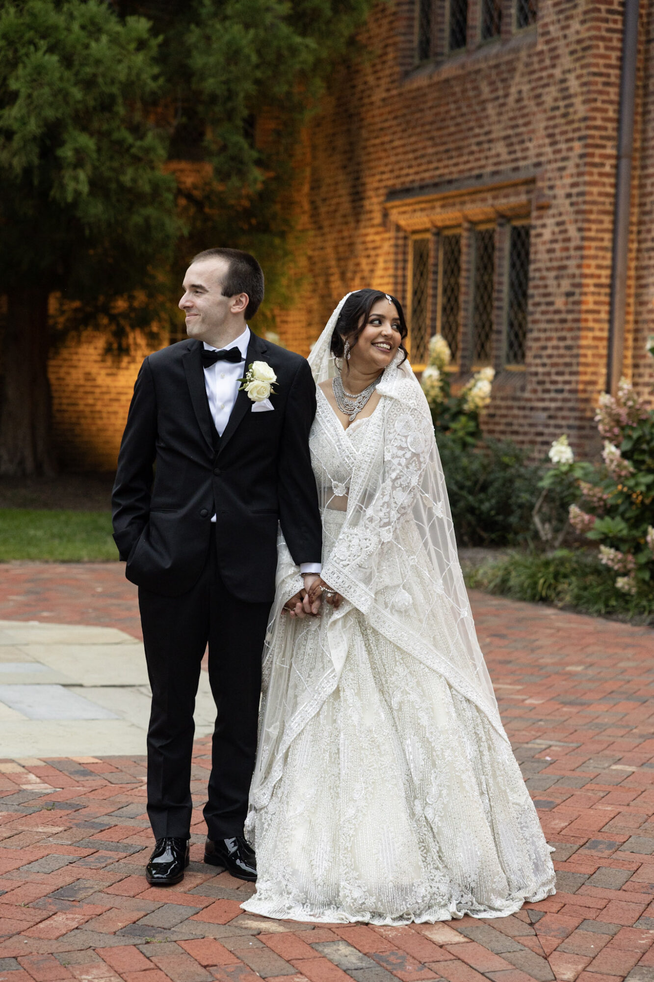 A bride and groom walk outdoors in front of a brick building, holding hands and smiling, dressed in wedding attire.