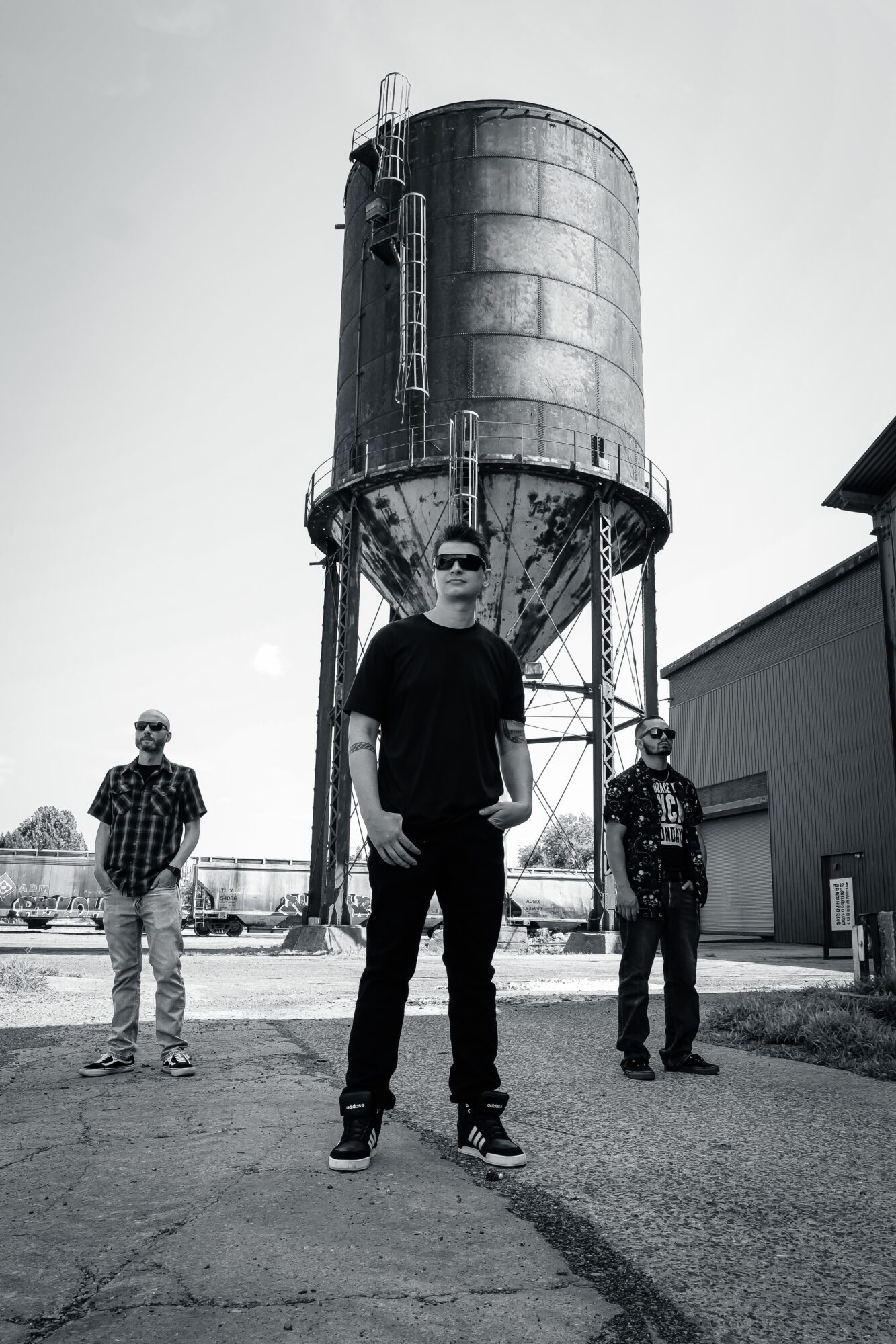 Three people standing outdoors in front of a large industrial tank structure, with a building on the right side.