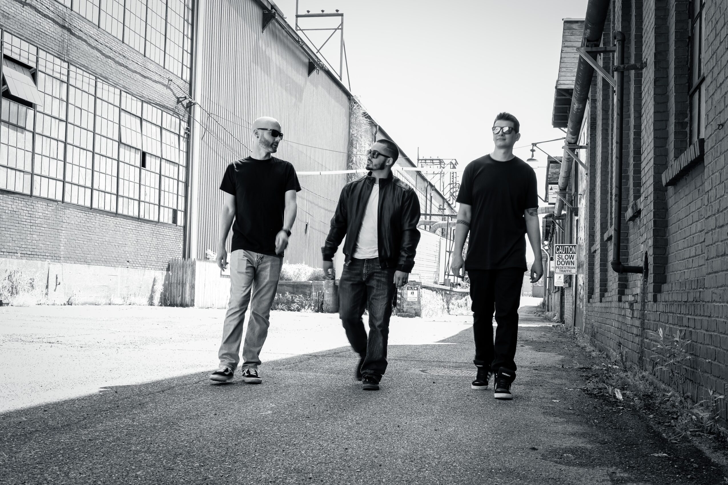 Three men walking down an urban alleyway, with industrial buildings on both sides, in black and white.