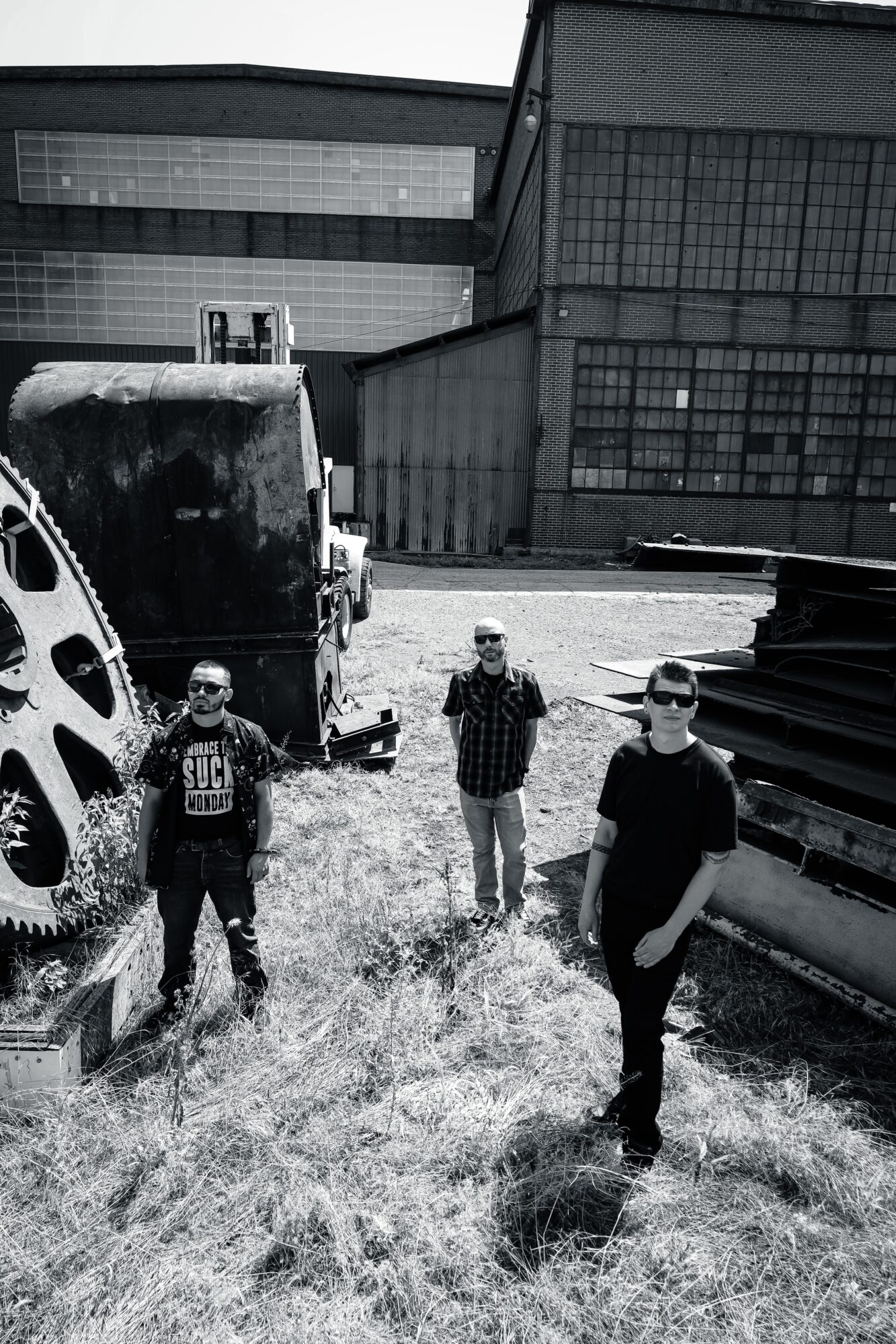 Four people standing outdoors near industrial buildings and machinery, with grass and a large wheel in the foreground.