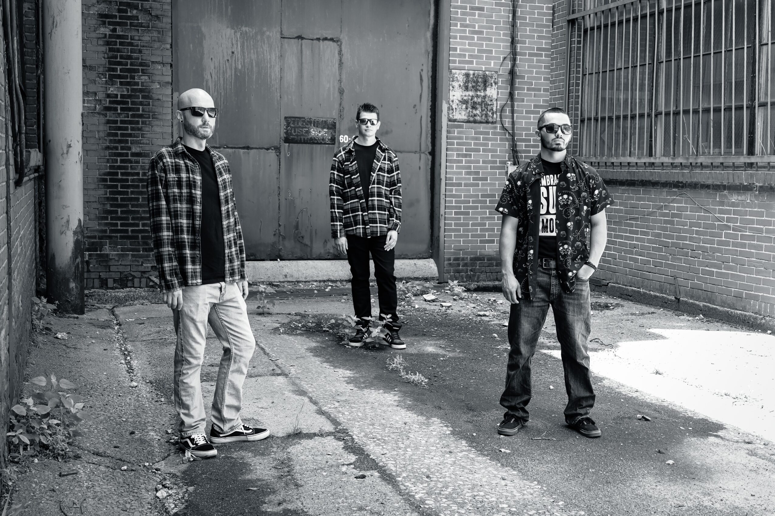 Three men standing in an alley with brick walls and a large metal door, black and white photo, casual clothing.