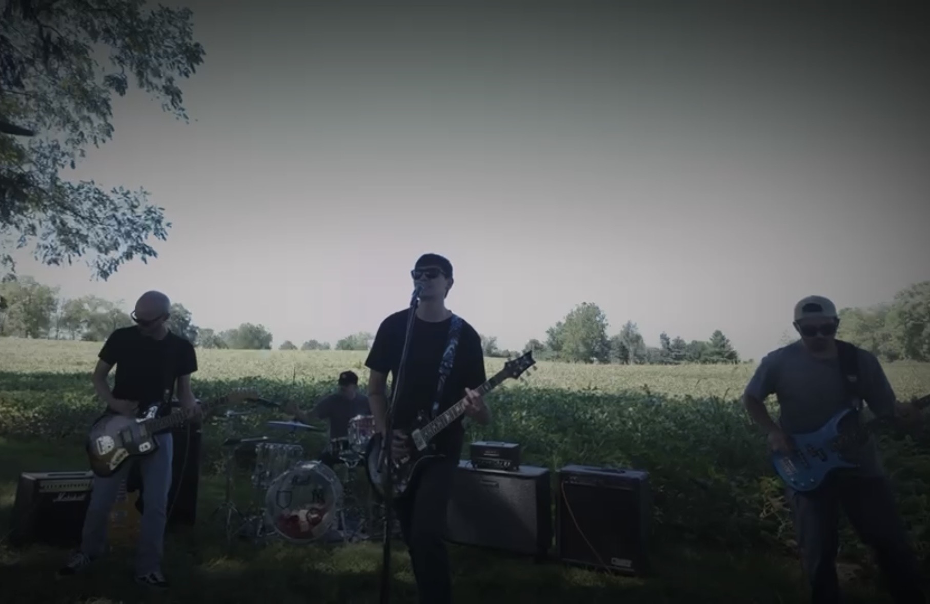 Band performing outdoors with three musicians playing guitars and drums, trees and open field in background.