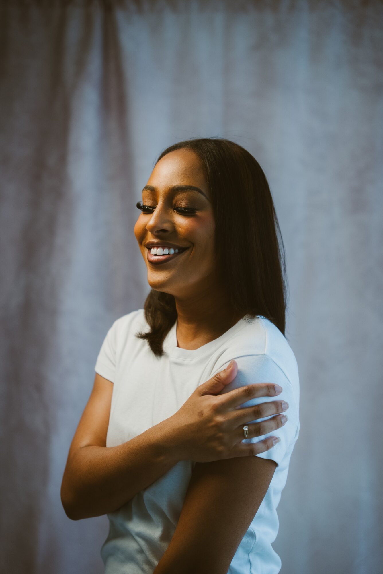 Smiling woman with shoulder-length dark hair wearing a white t-shirt, standing against a plain background.
