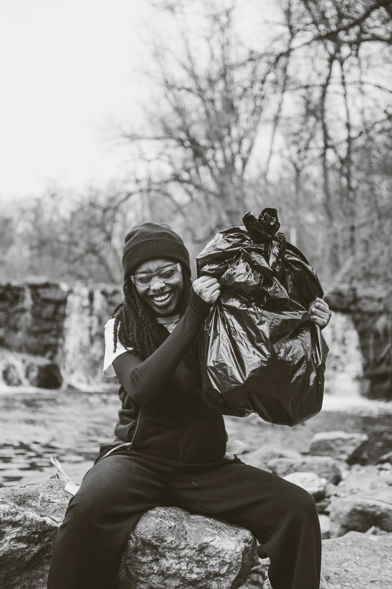 Person sitting on a rock holding a large bag outdoors, smiling, with trees and a waterfall in the background.
