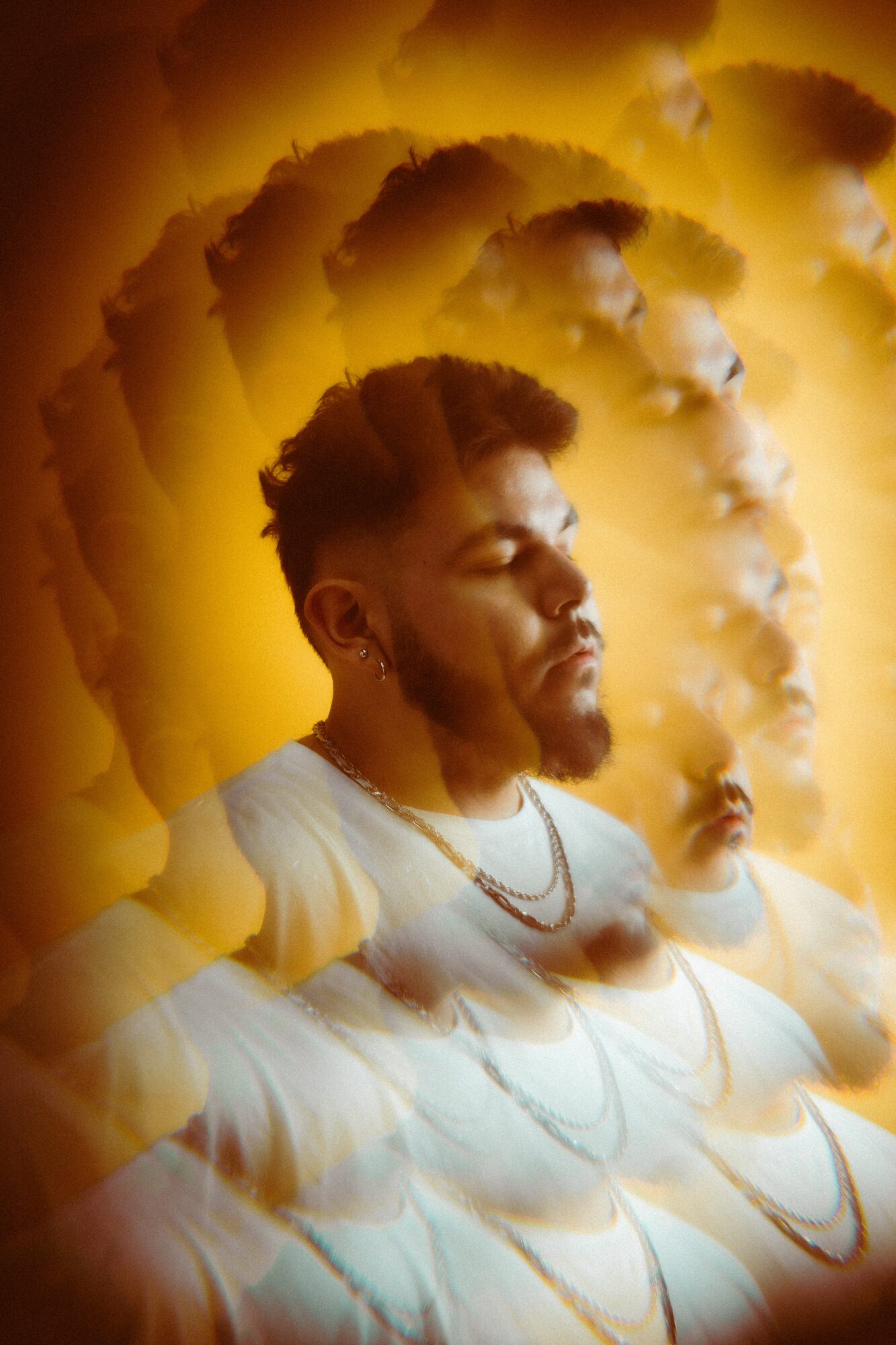 Person with eyes closed, multiple exposure effect, warm yellow background, wearing a white shirt and jewelry.