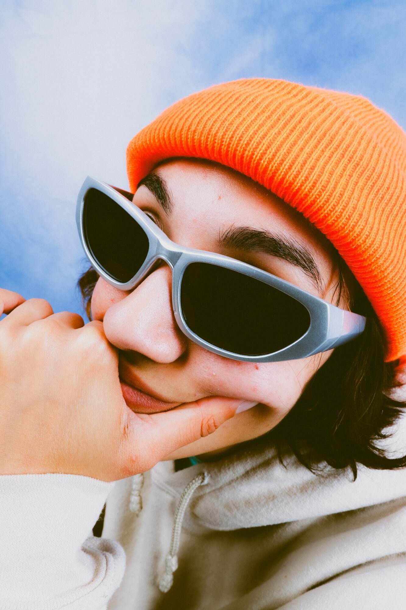 Person wearing orange beanie and sunglasses, resting chin on hand, outdoors with blue sky background.