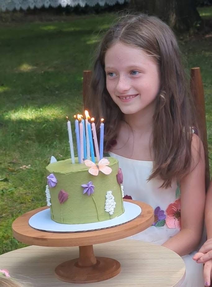 Young girl with long hair smiling at a birthday cake outdoors, decorated with butterflies and purple flowers.