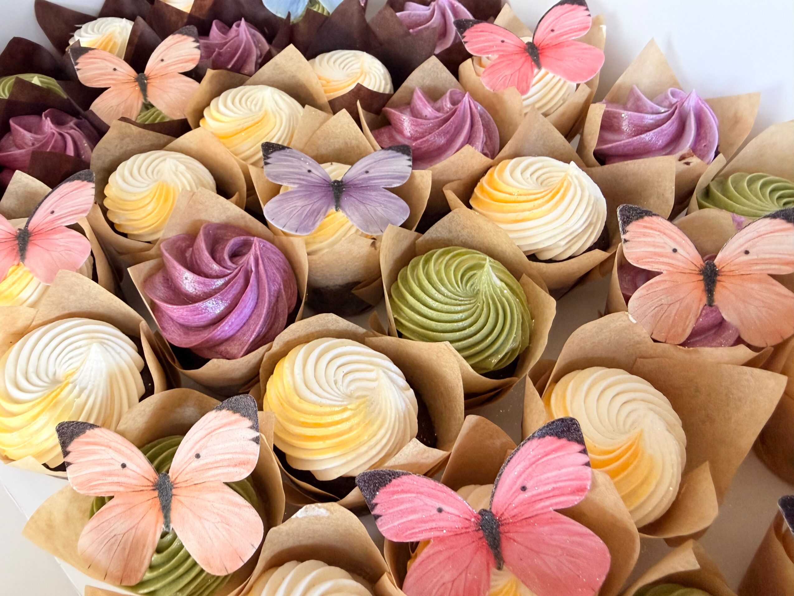 Assorted colorful cupcakes decorated with butterfly-shaped toppers, arranged in paper wrappers, with swirled frosting in various colors.