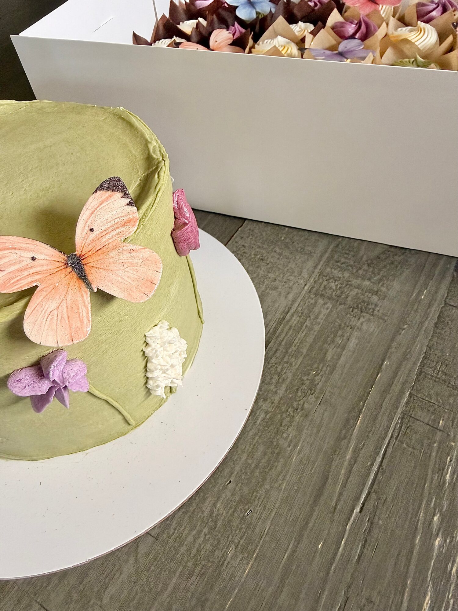 Green cake with butterfly and flower decorations on a white cake stand, with a box of colorful flowers in the background.