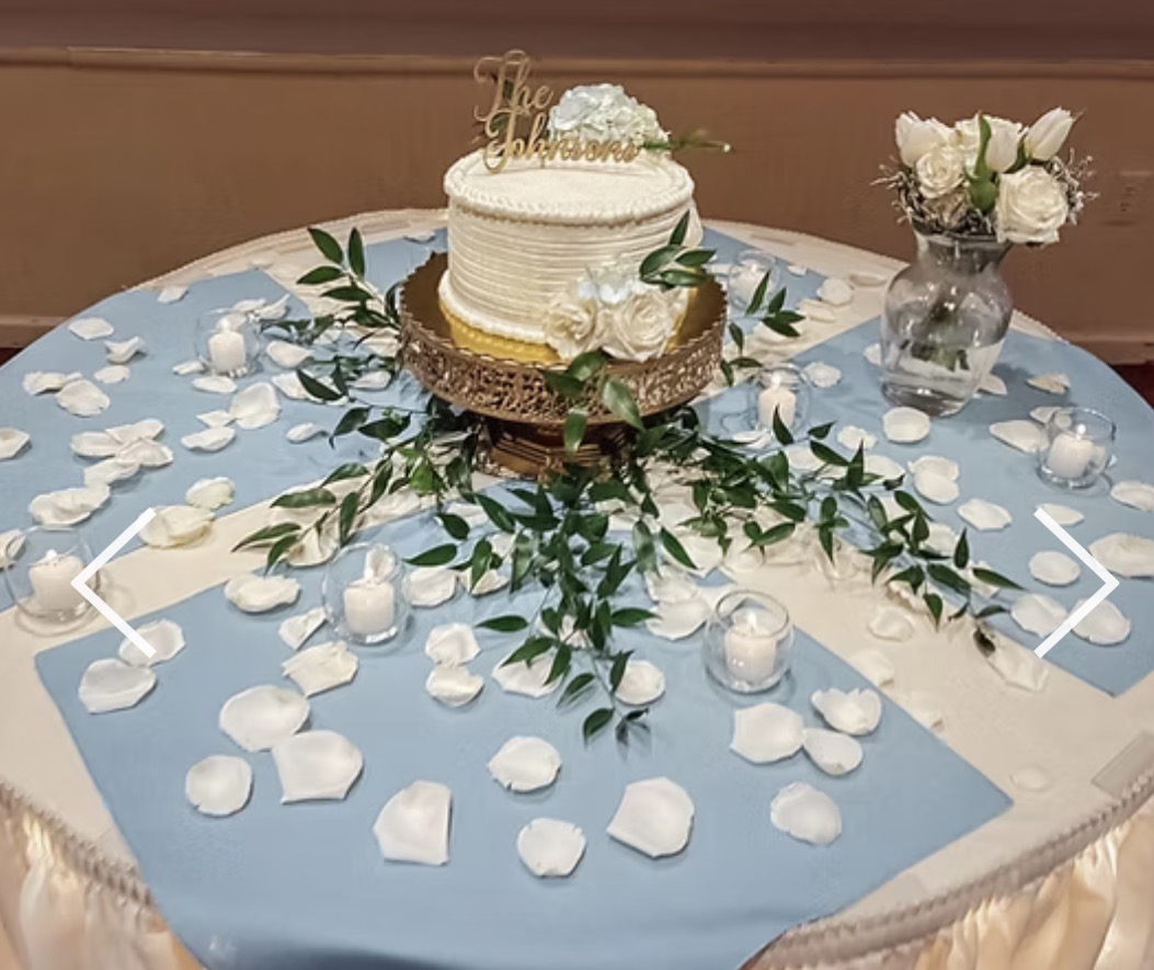 Round wedding cake with white frosting, gold topper, surrounded by white rose bouquet and scattered rose petals on table