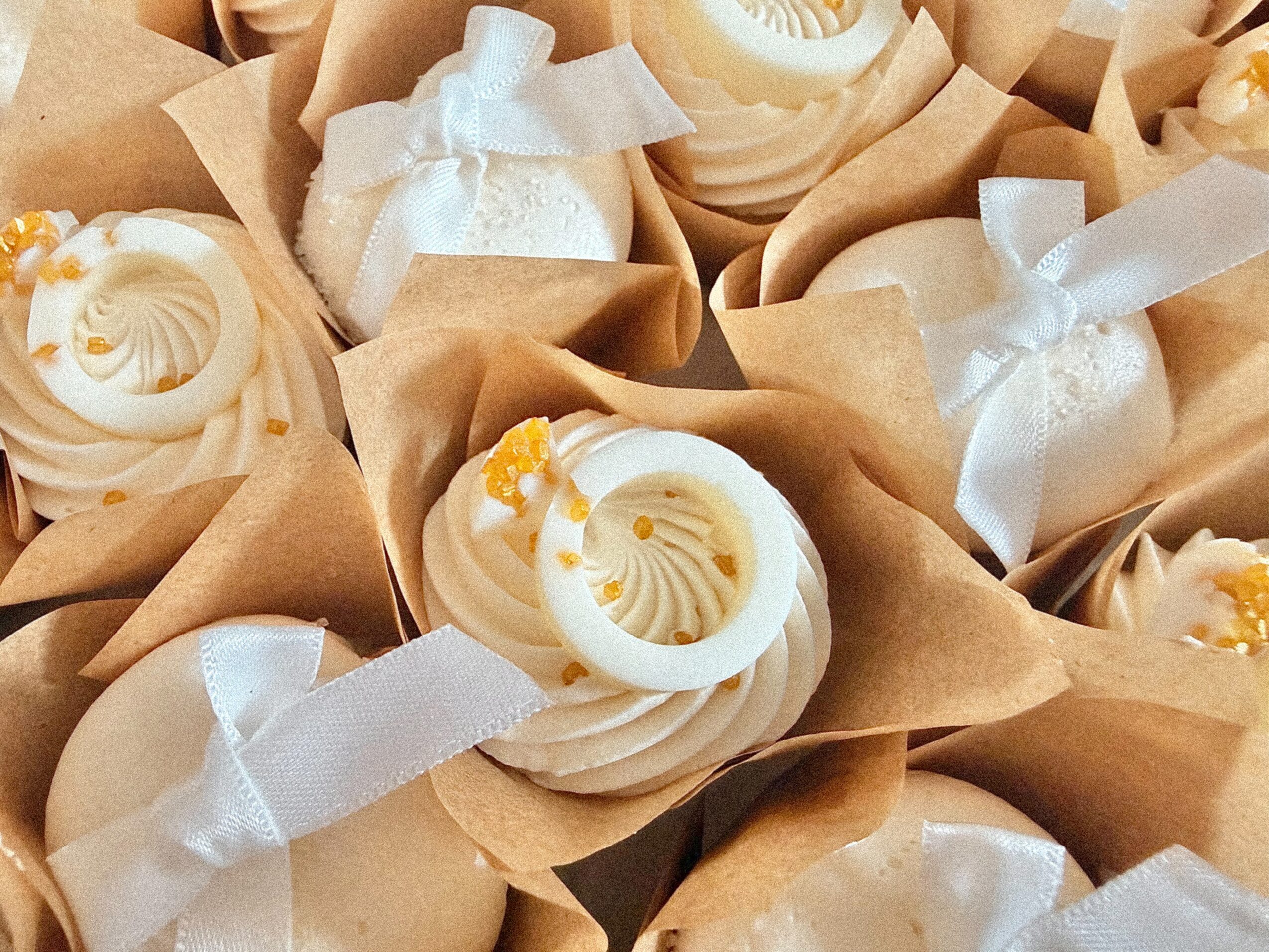 Cupcakes decorated with white frosting, yellow sprinkles, and white ribbons, arranged in paper wrappers.