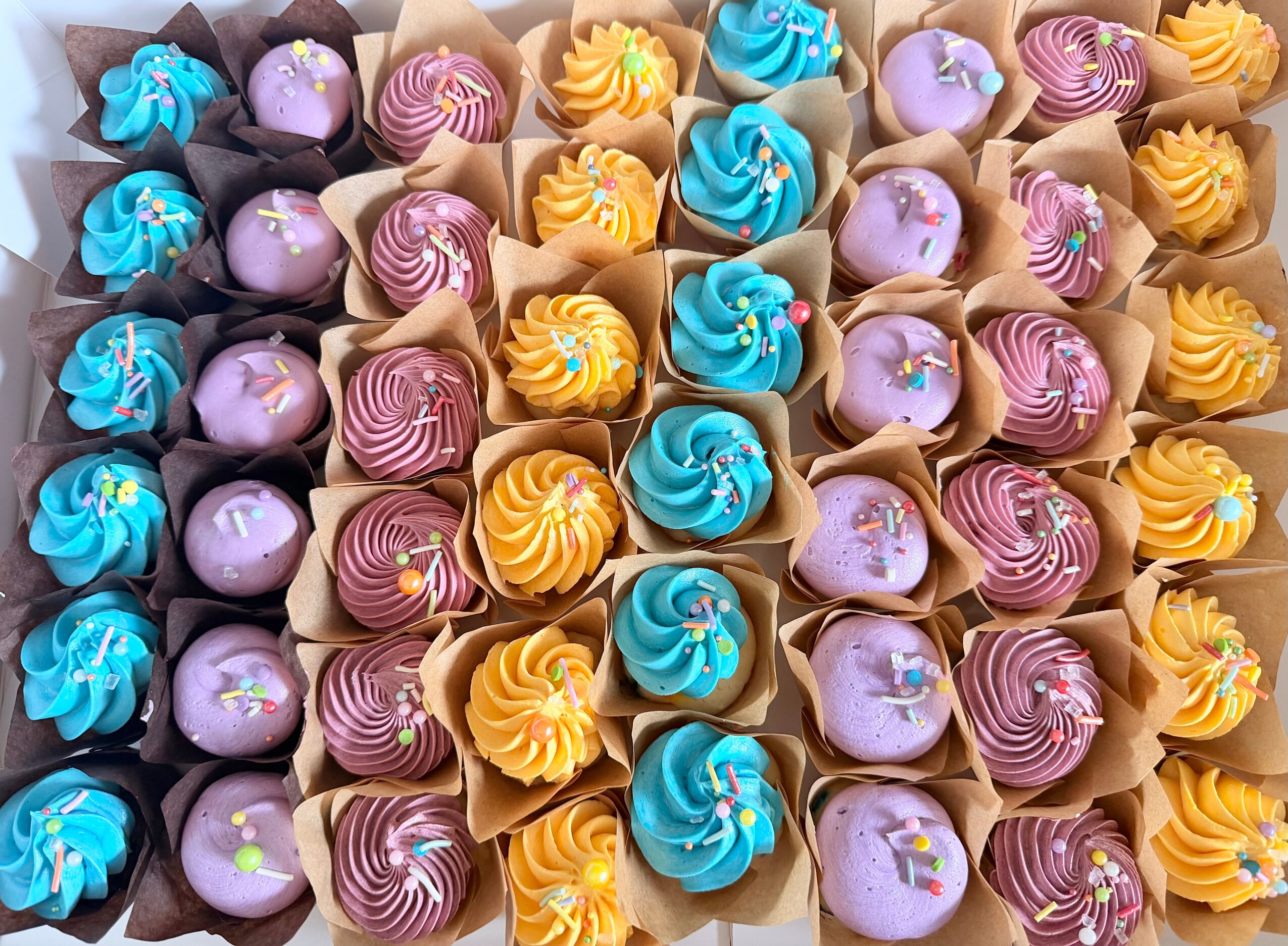 Colorful cupcakes with blue, purple, and yellow frosting arranged in rows.