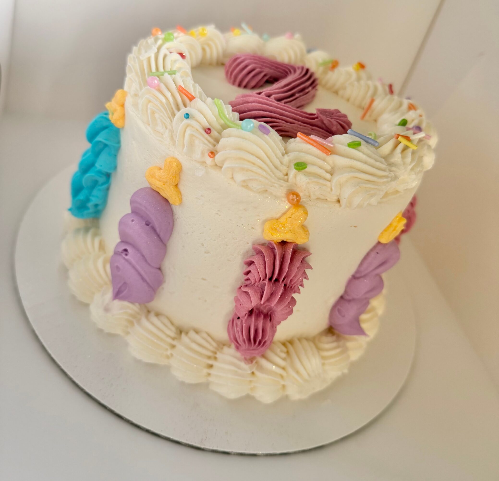Round birthday cake with white frosting, decorated with colorful piped icing, sprinkles, and small yellow and purple figures, on a white surface.