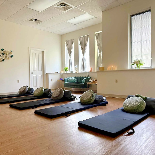 Yoga mats with bolsters and blankets arranged in a spacious, well-lit room with windows and a green sofa.