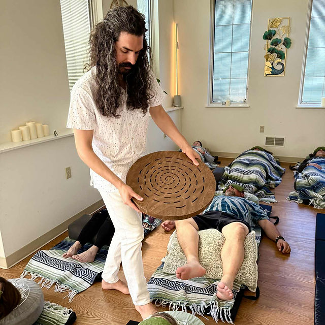 Person holding a circular wooden object in a room with people lying on cushions, windows, and wall art.
